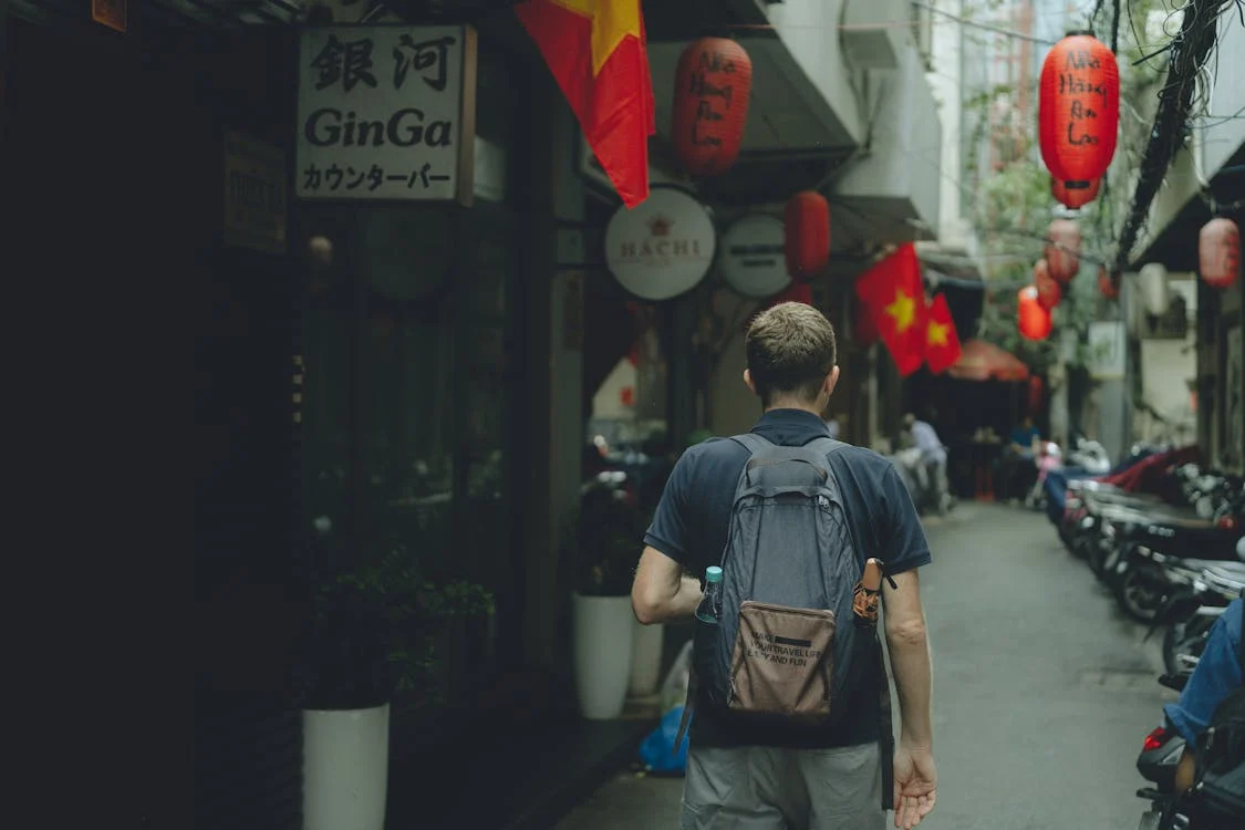 A man walking down a narrow alleyway with red lanterns hanging from the ceiling / Credits: Khanh Nguyen (Pexels)